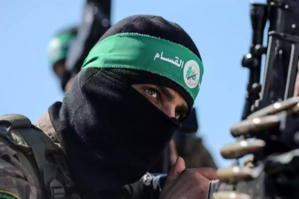 Persona con pasamontañas negro y cinta verde en la cabeza con letras árabes, sosteniendo un arma Persona con pasamontañas negro y cinta verde en la cabeza con letras árabes, sosteniendo un arma