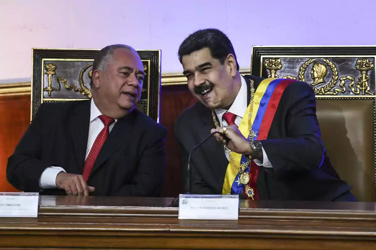 Dos hombres en trajes formales conversan y sonríen mientras están sentados en sillas ornamentadas en un entorno oficial.