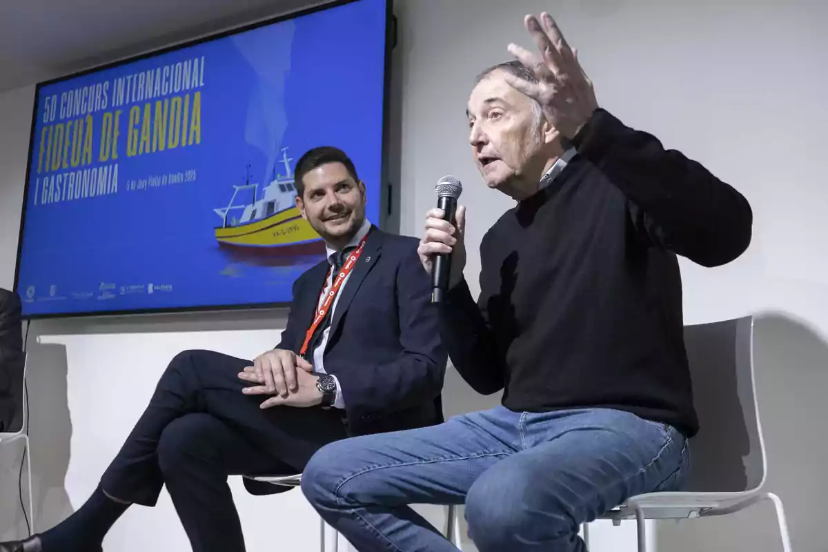 Dos hombres sentados en sillas, uno de ellos hablando con un micrófono en la mano, frente a una pantalla que anuncia el 50 Concurso Internacional de Fideuà de Gandia y Gastronomía.