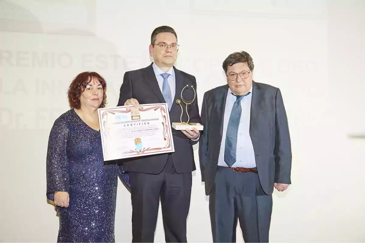 Tres personas posan para una foto en un evento de premiación, una de ellas sostiene un certificado y un trofeo.