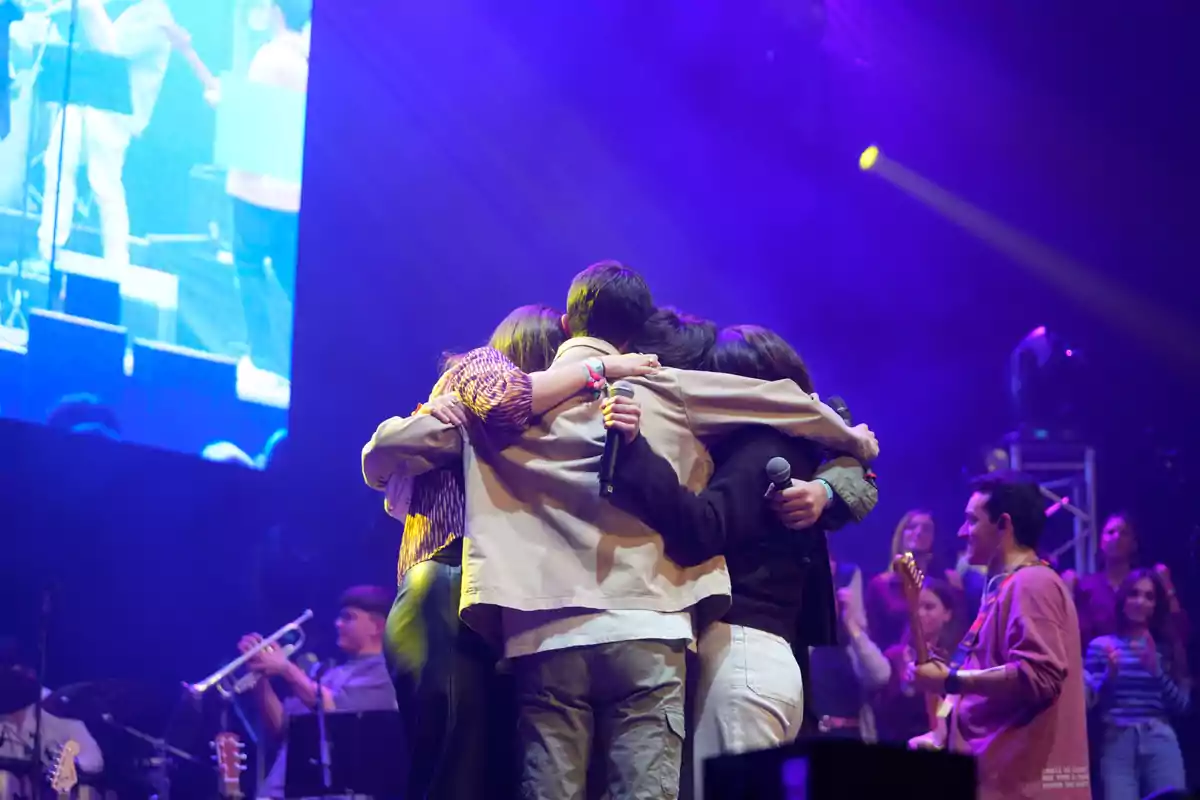 Un grupo de personas se abraza en el escenario durante un concierto, con luces de colores y músicos tocando instrumentos al fondo.