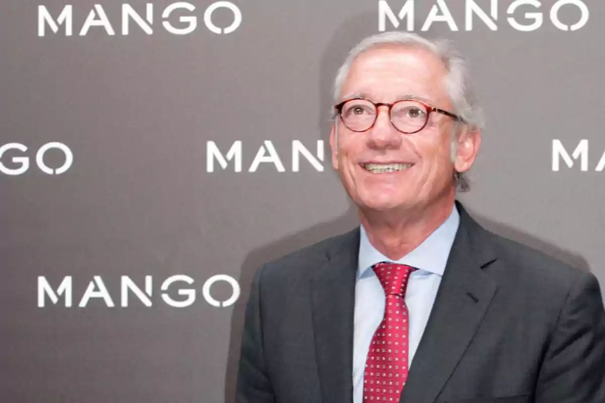 A man with glasses and a suit smiles in front of a background with the Mango logo.