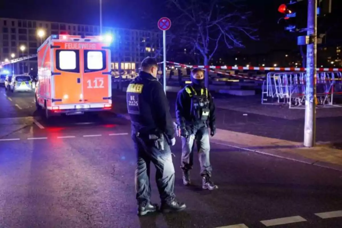 Dos personas con uniformes de policía están de pie en una calle iluminada por luces de emergencia, junto a un vehículo de bomberos de color naranja.