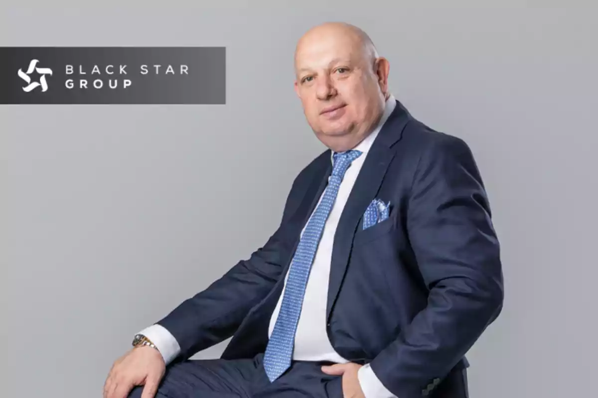 A bald man in a blue suit and light blue tie is sitting in front of a gray background, next to a logo that says "Black Star Group".