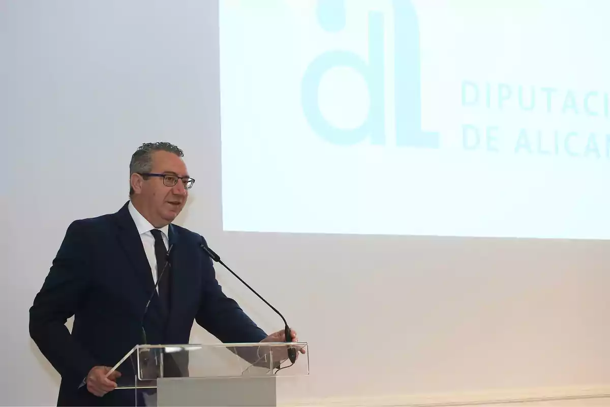 A man in a suit speaking at a podium with the logo of the Legislature de Alicante in the background.