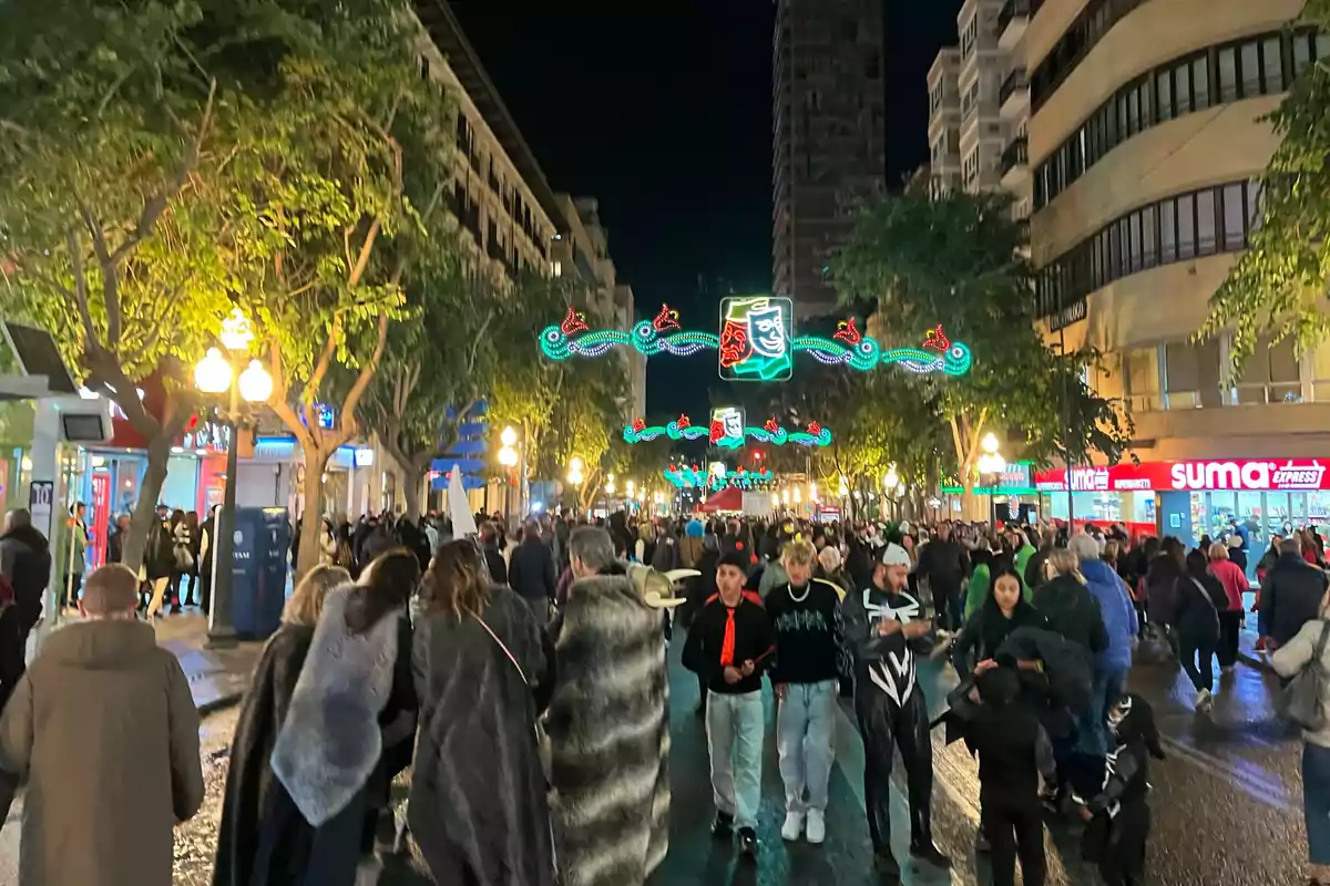 A crowd of people walks down a street lit with decorative lights and trees on the sides during the night.