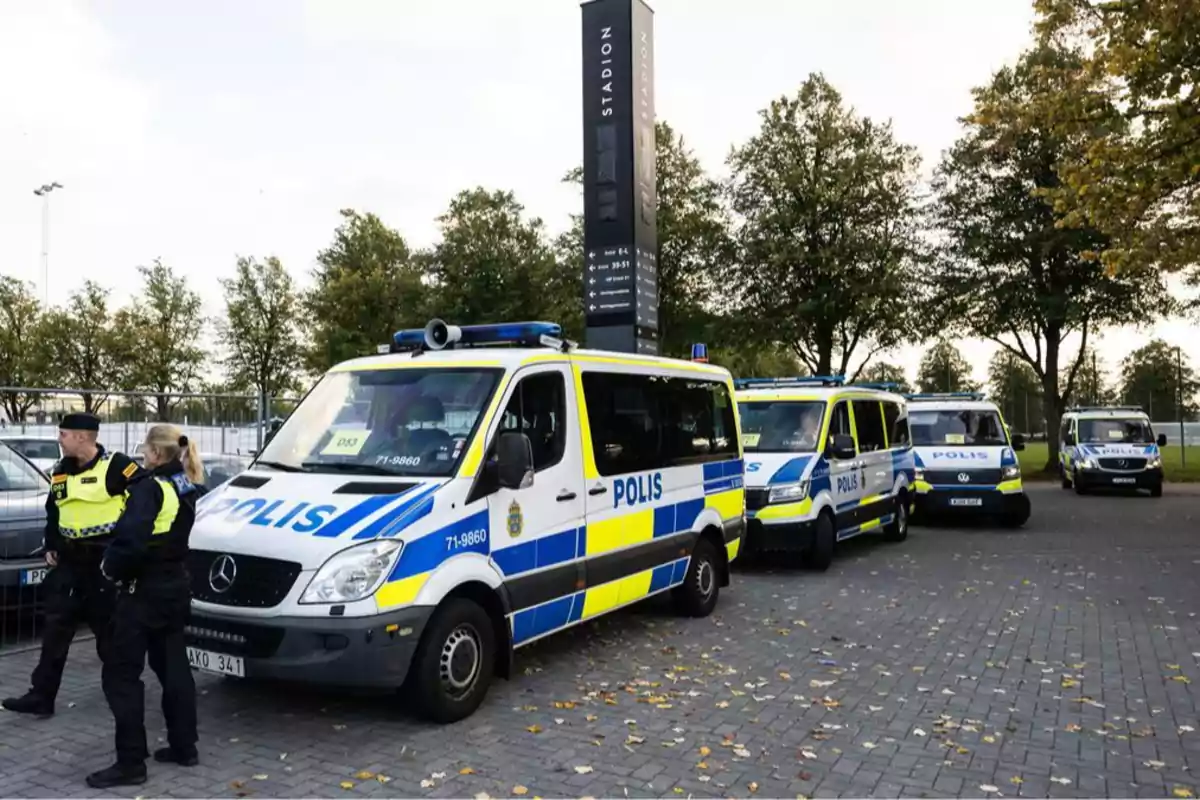 Varios vehículos policiales estacionados en fila junto a un grupo de oficiales en un área al aire libre con árboles y un letrero alto.