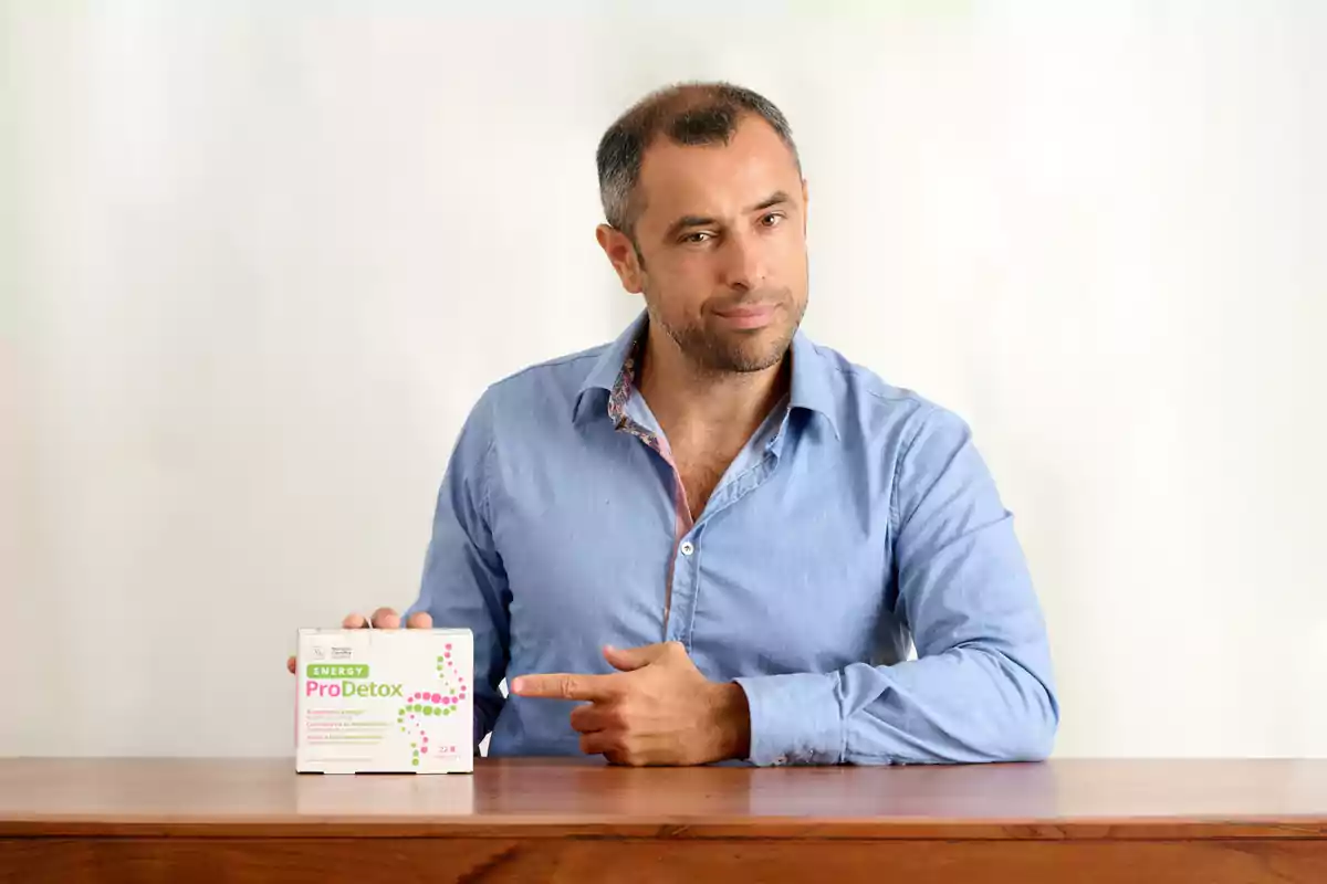 Hombre con camisa azul señalando una caja de suplemento ProDetox sobre una mesa de madera.