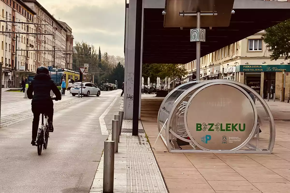 Una persona en bicicleta circula por una calle urbana con un aparcamiento para bicicletas a la derecha y edificios al fondo.