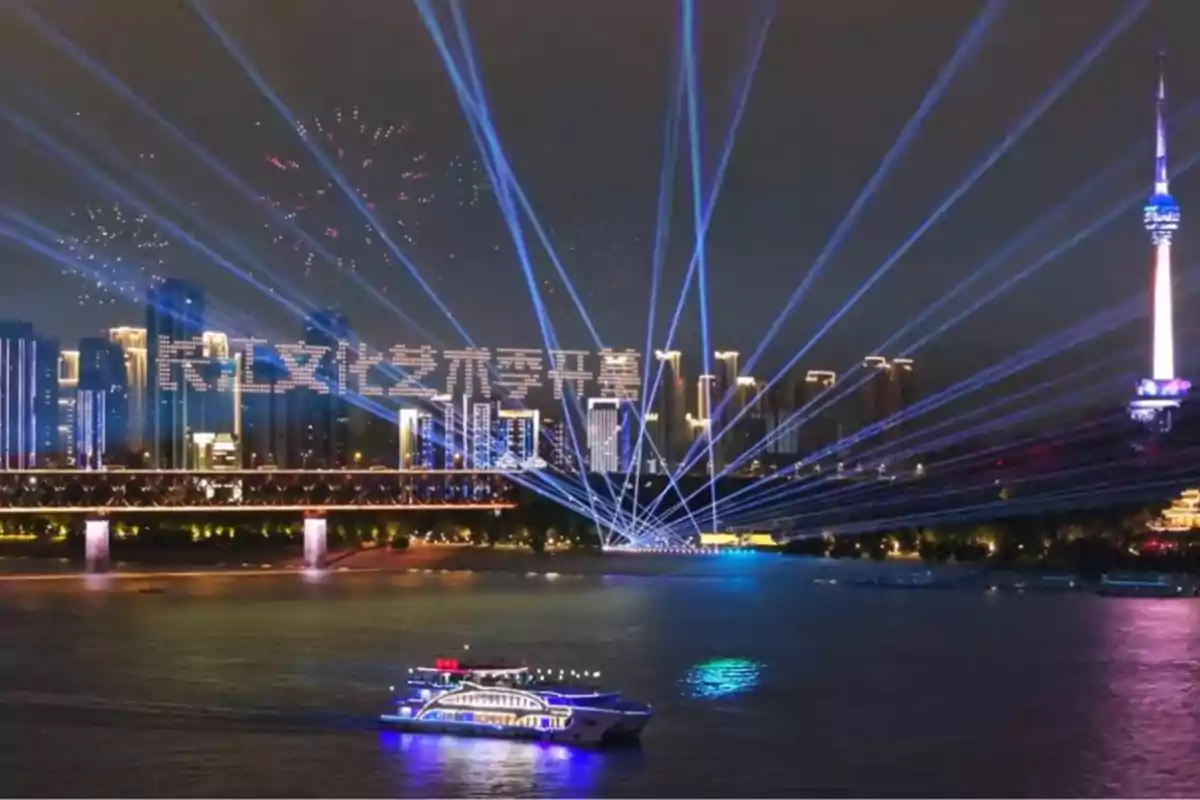 Un espectáculo de luces y fuegos artificiales ilumina el cielo nocturno sobre una ciudad con rascacielos, un puente y un barco en el río.