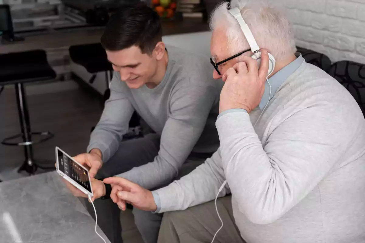 Un hombre joven y un hombre mayor están sentados juntos, mirando una tableta mientras el hombre mayor usa auriculares.