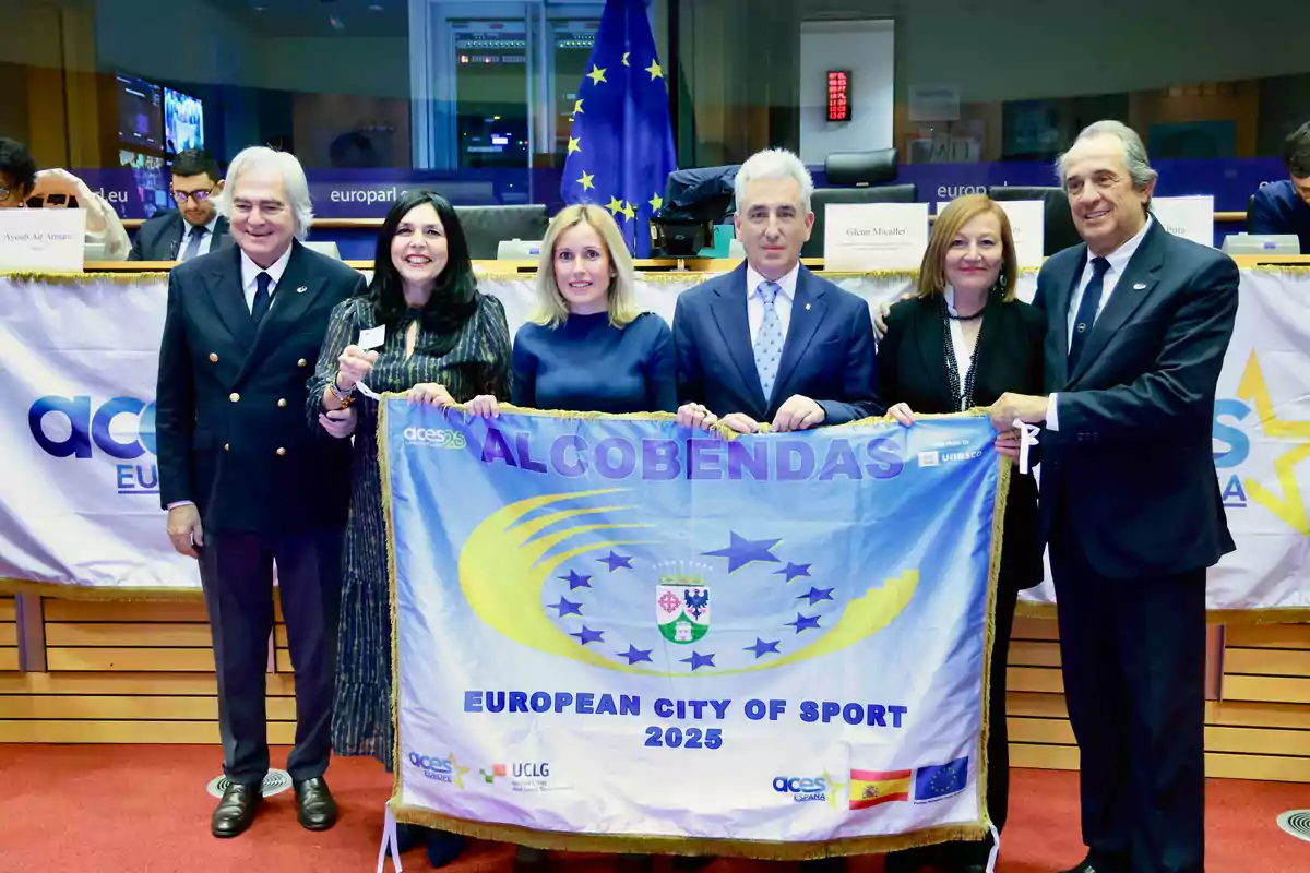 Un grupo de personas sostiene una bandera que dice "Alcobendas European City of Sport 2025" en un evento oficial con banderas de la Unión Europea al fondo.