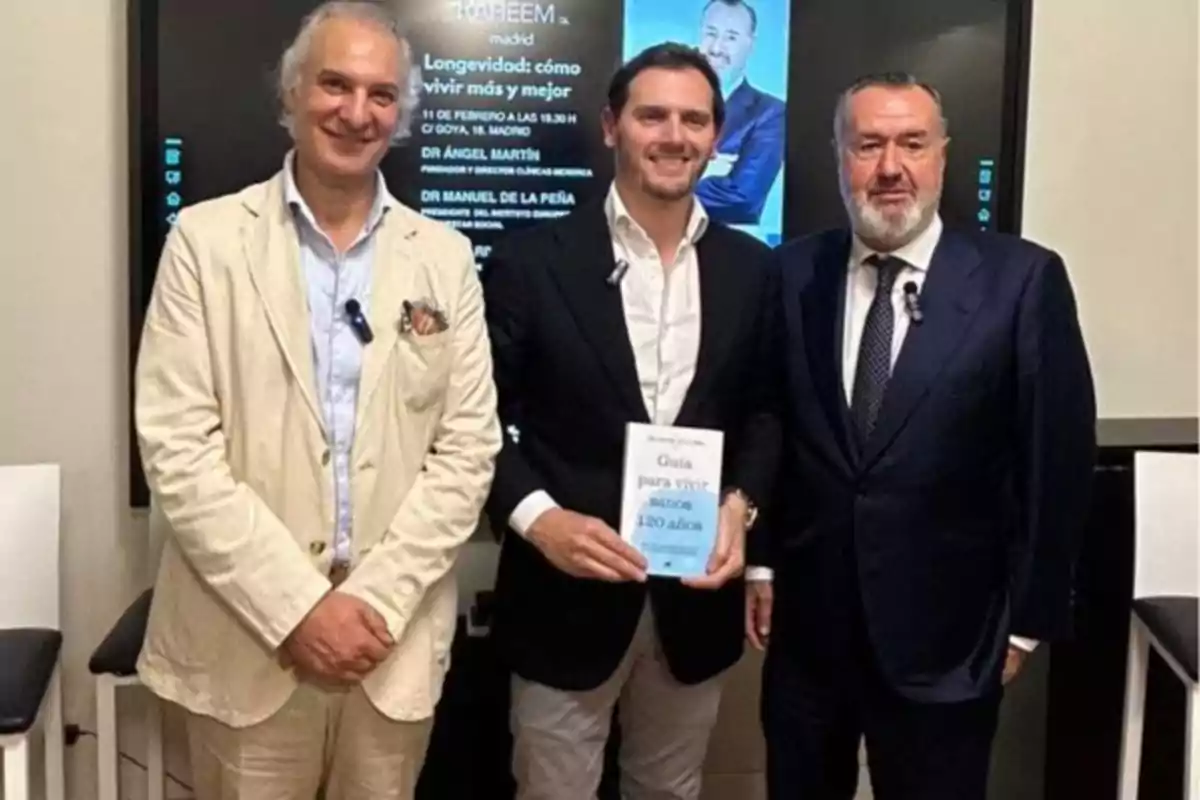 Tres hombres posan juntos en un evento, uno de ellos sostiene un libro titulado "Guía para vivir 120 años".