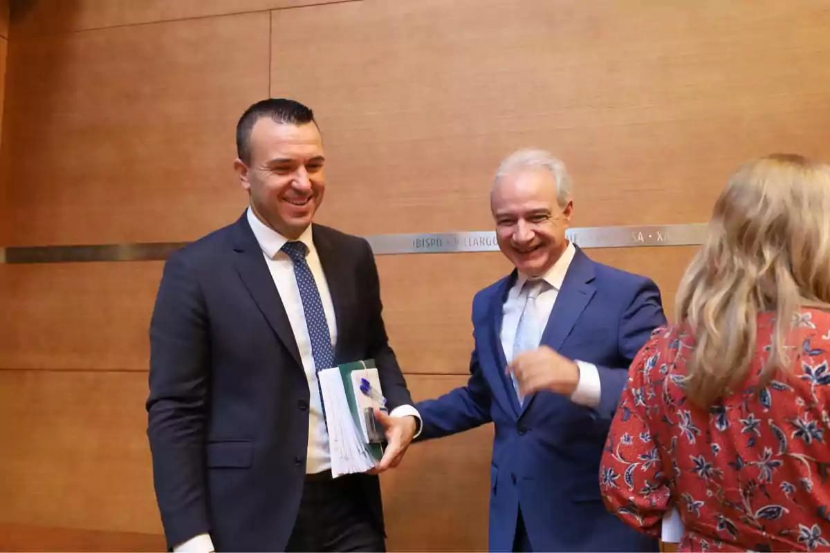 Two men in suits smile while conversing in a formal setting, one of them holding documents.