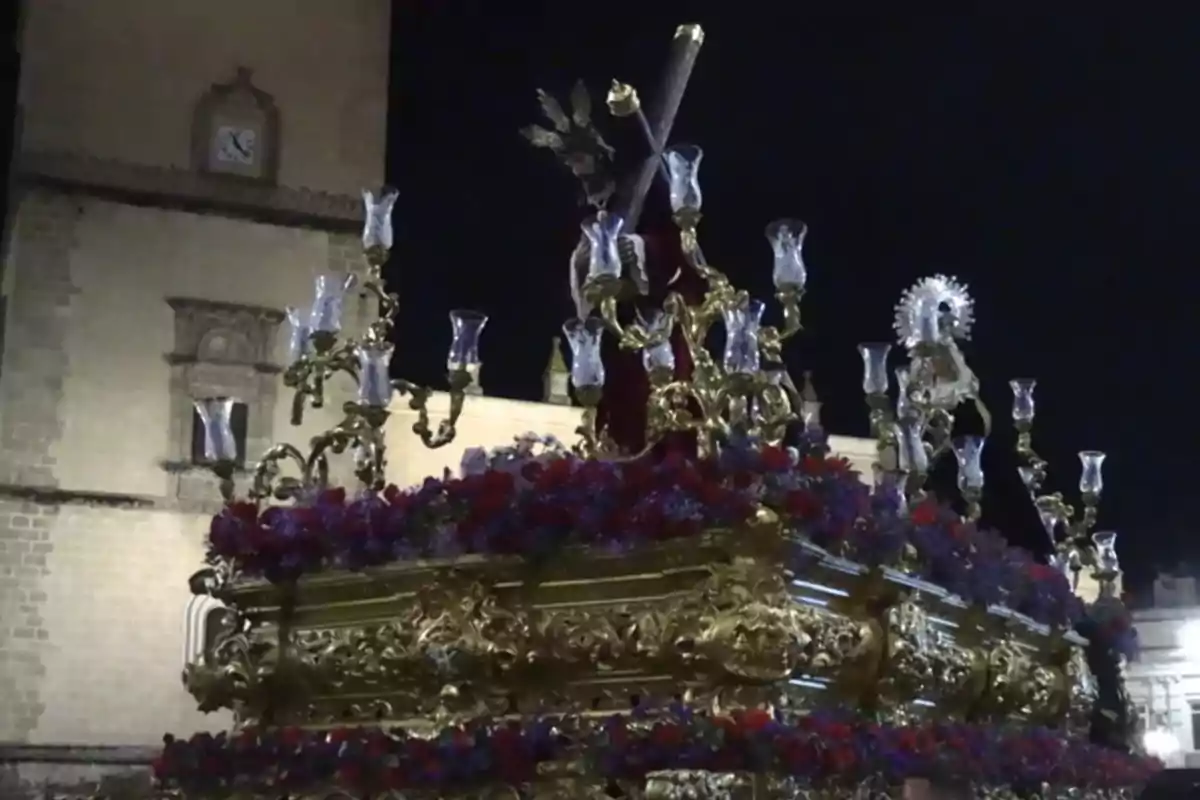 Una imagen de un paso de Semana Santa adornado con flores y candelabros, con una figura religiosa en el centro, frente a un edificio histórico iluminado por la noche. Una imagen de un paso de Semana Santa adornado con flores y candelabros, con una figura religiosa en el centro, frente a un edificio histórico iluminado por la noche.