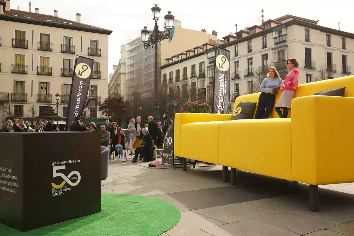 Dos mujeres están sentadas en un sofá amarillo gigante en una plaza rodeada de edificios mientras varias personas observan el evento