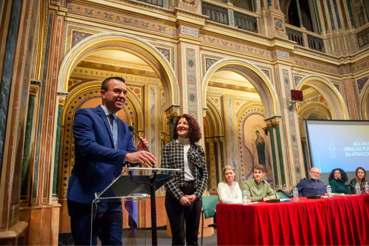 A man speaking at a podium in a room decorated with arches and golden details, joined by a woman and several people seated at a table with a red tablecloth.