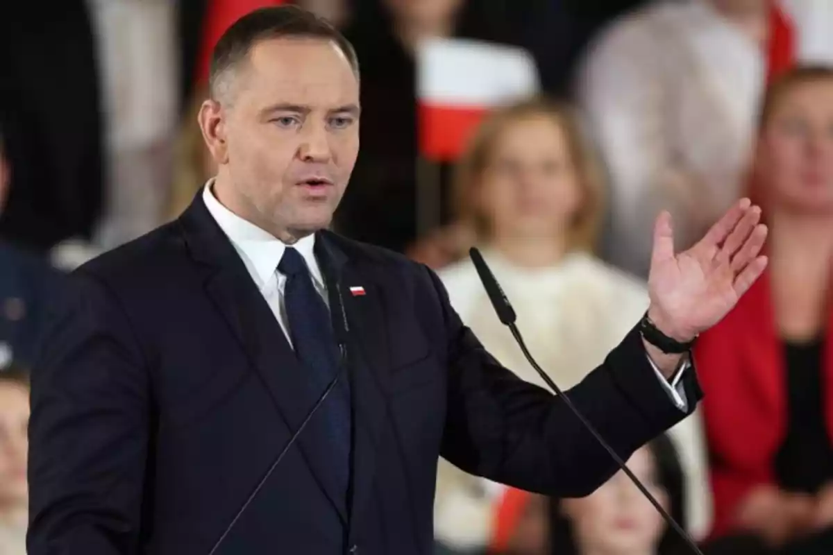 Hombre de traje oscuro hablando en un podio con micrófonos y personas desenfocadas al fondo sosteniendo banderas Hombre de traje oscuro hablando en un podio con micrófonos y personas desenfocadas al fondo sosteniendo banderas
