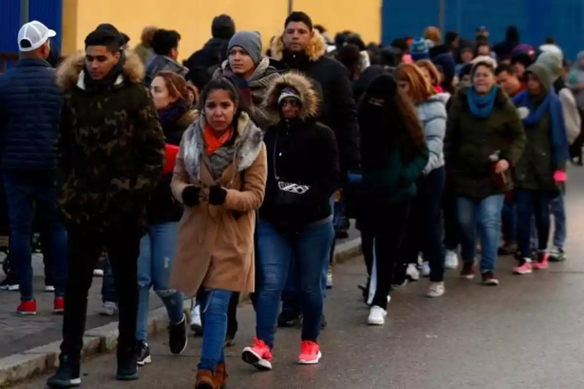Un grupo de personas camina en fila por una acera, abrigadas con chaquetas y bufandas en un día frío.