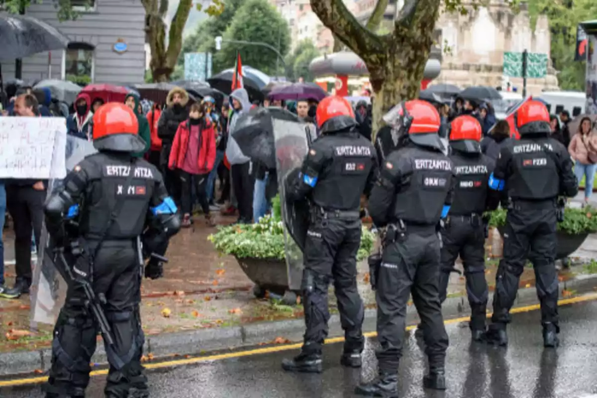 Police officers with red helmets and riot gear watch a group of protesters in the rain.