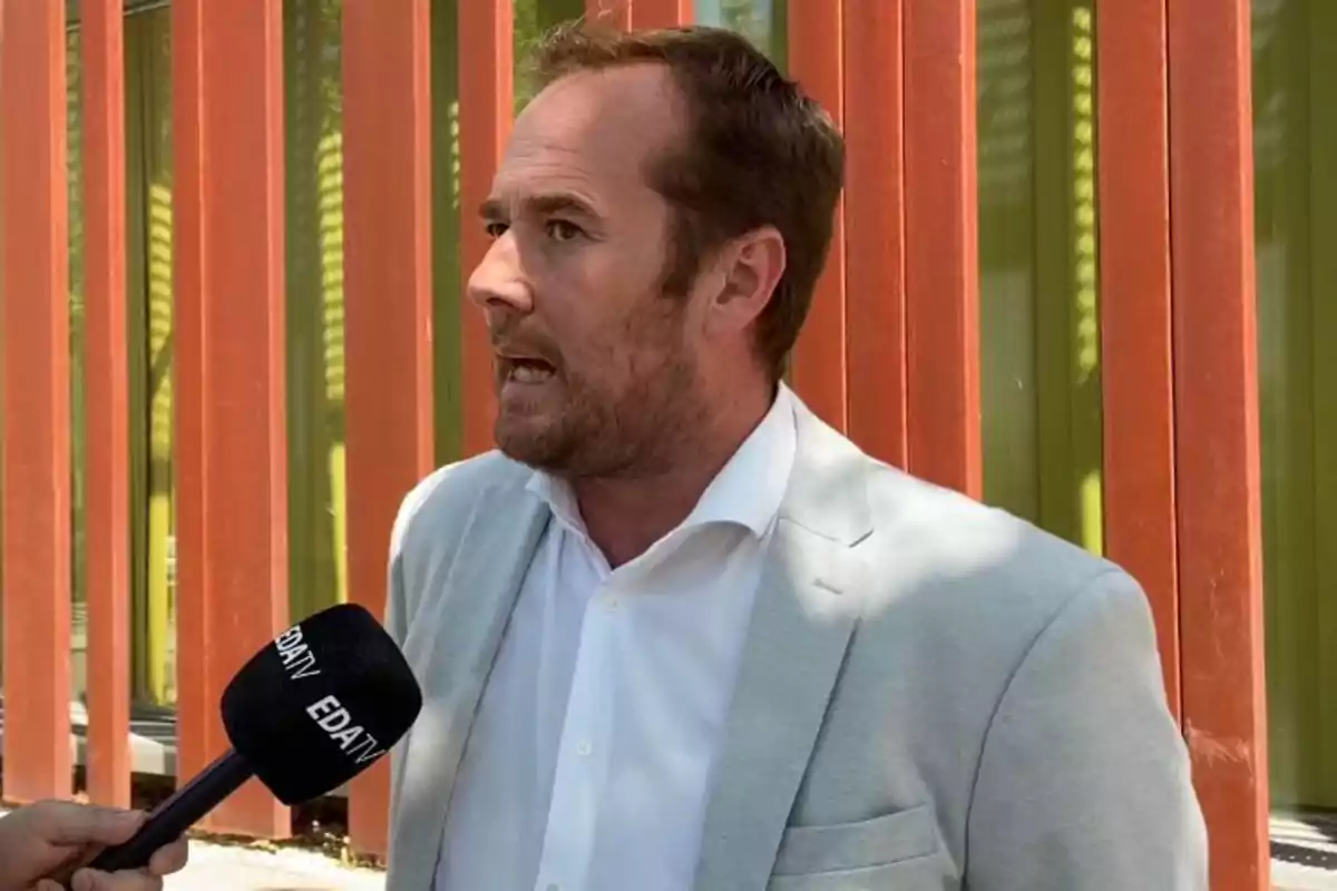 Hombre con saco claro y camisa blanca siendo entrevistado por un micrófono de EDATV frente a una pared con paneles naranjas y verdes. Hombre con saco claro y camisa blanca siendo entrevistado por un micrófono de EDATV frente a una pared con paneles naranjas y verdes.