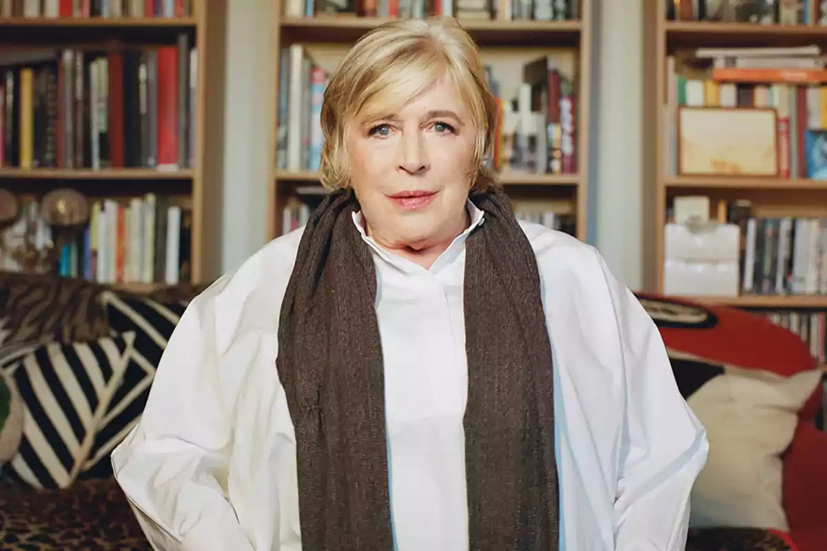 A person with blonde hair and a dark scarf is standing in front of a bookshelf full of books.