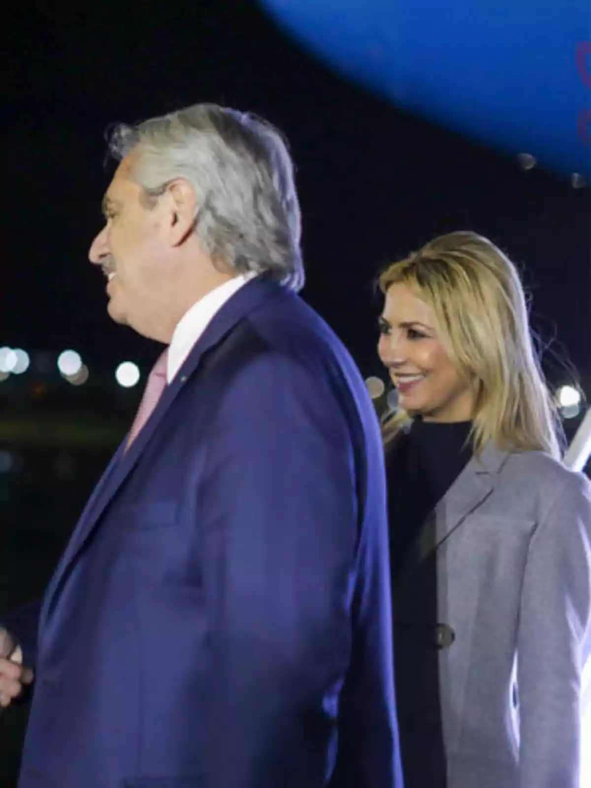 Dos personas sonrientes, un hombre de cabello canoso y una mujer rubia, ambos vestidos formalmente, en un entorno nocturno con luces de fondo.