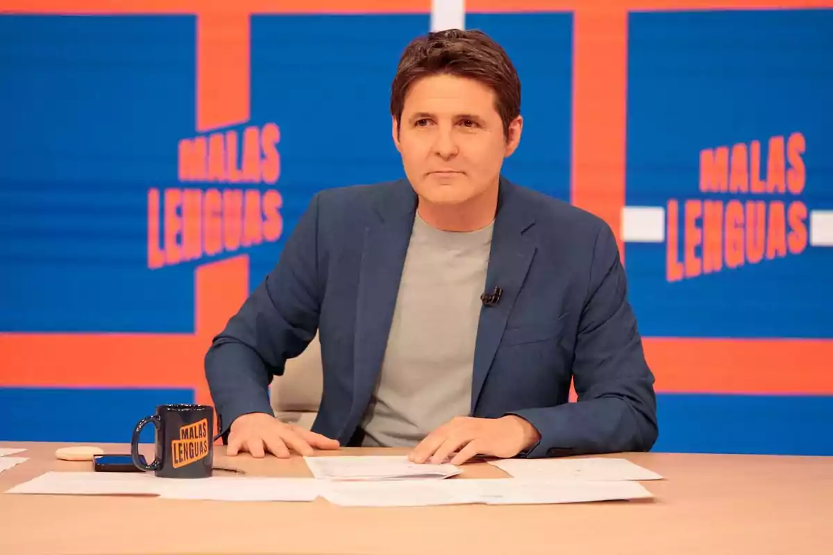 Un hombre sentado en un set de televisión con un fondo azul y naranja que dice Un hombre sentado en un set de televisión con un fondo azul y naranja que dice