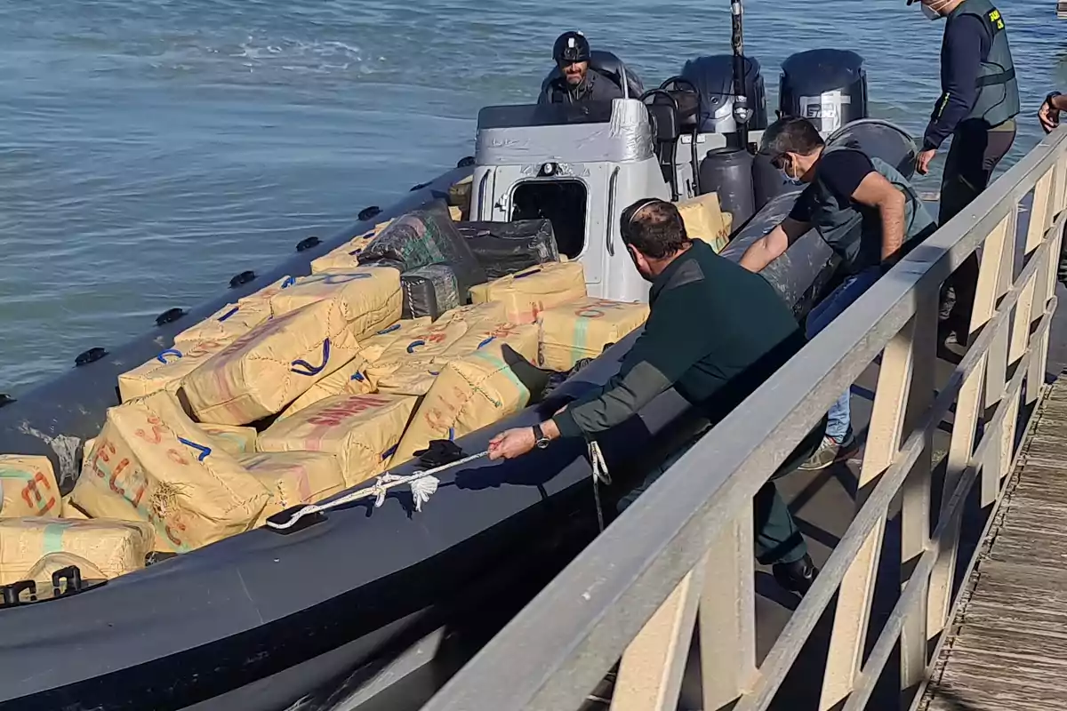 A group of people on a dock inspects a boat loaded with numerous wrapped packages.