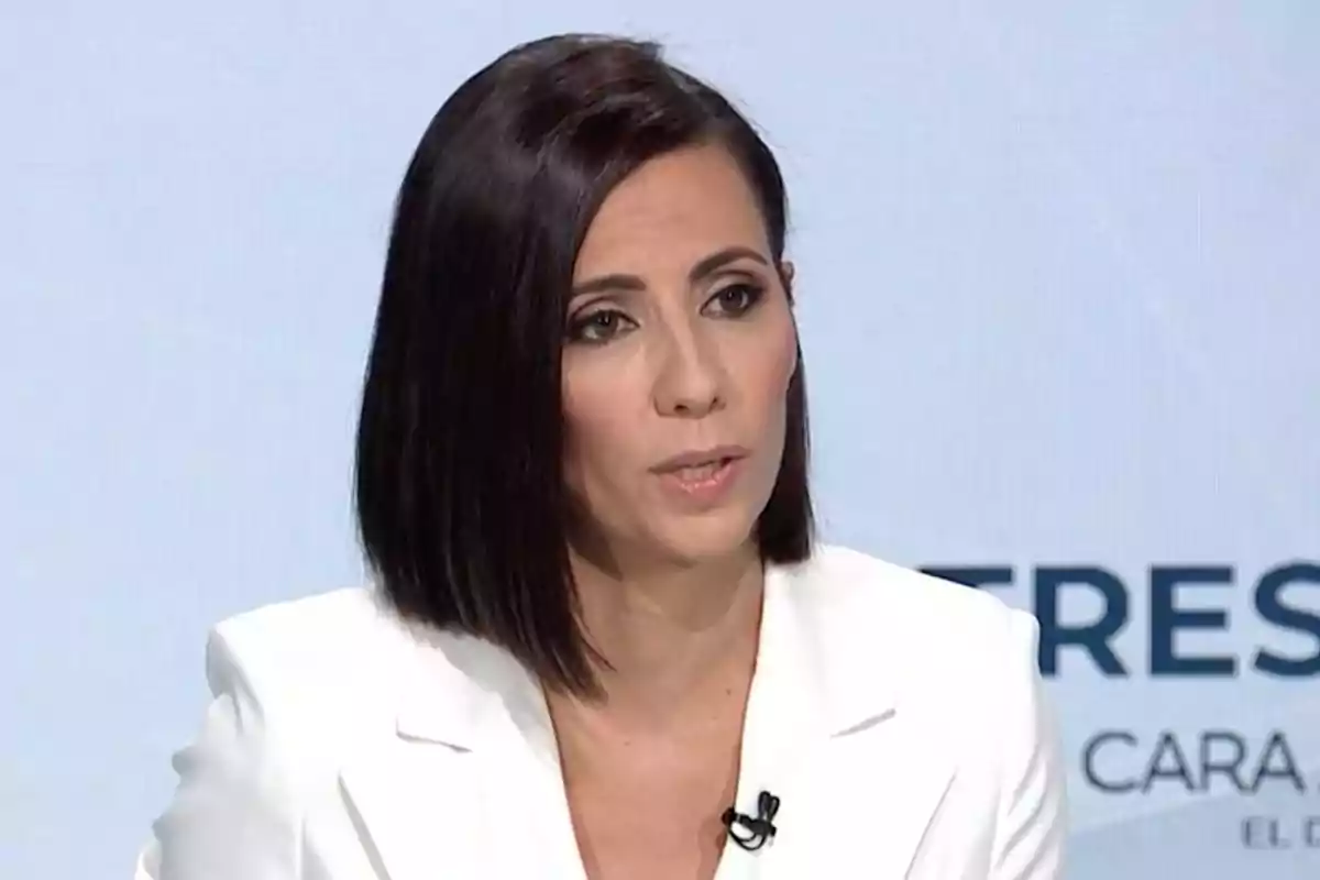 Una mujer con cabello oscuro y lacio lleva un traje blanco y está hablando en un entorno de estudio de televisión.