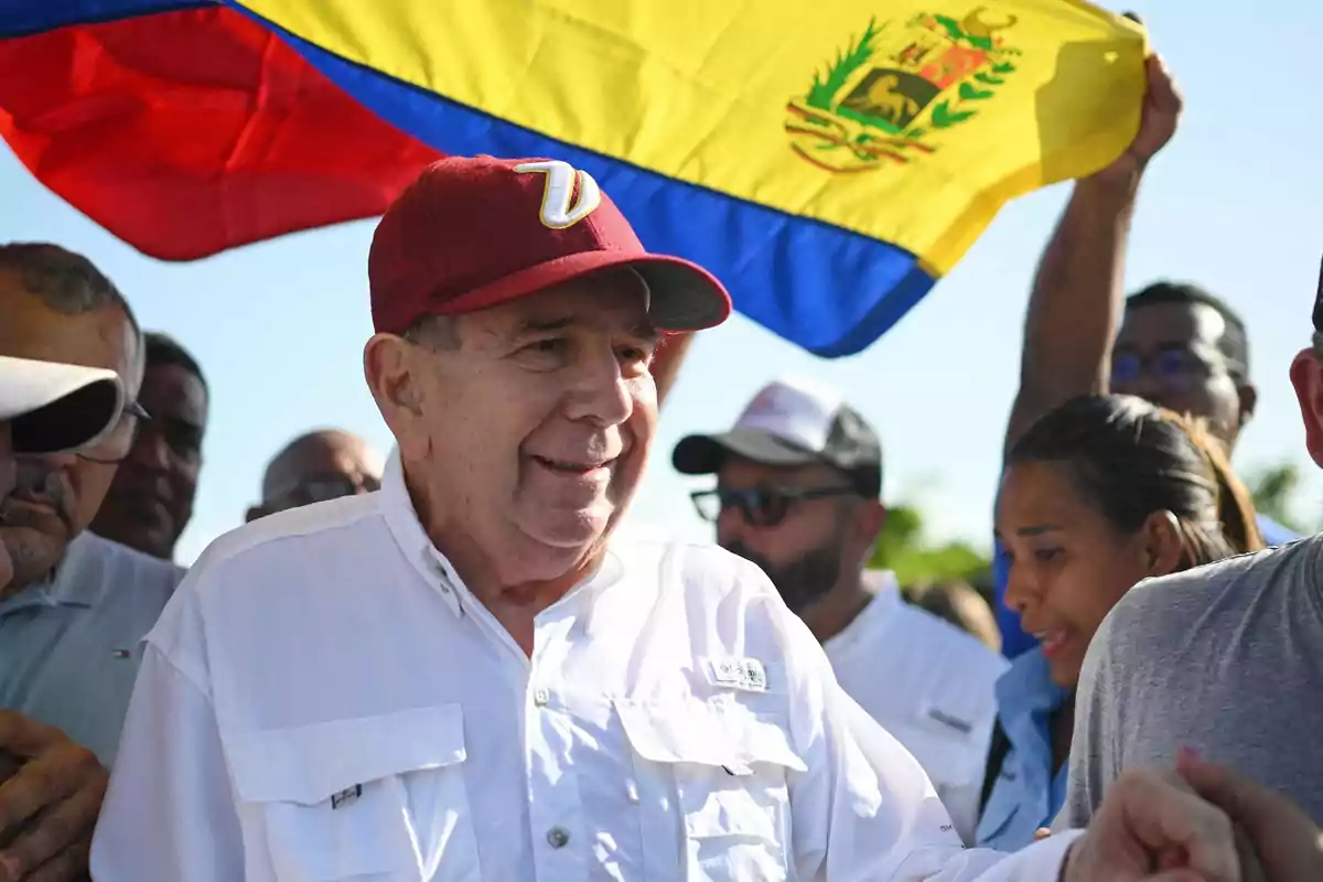 Un hombre mayor con una gorra roja y una camisa blanca está rodeado de personas mientras una bandera de Venezuela ondea en el fondo.