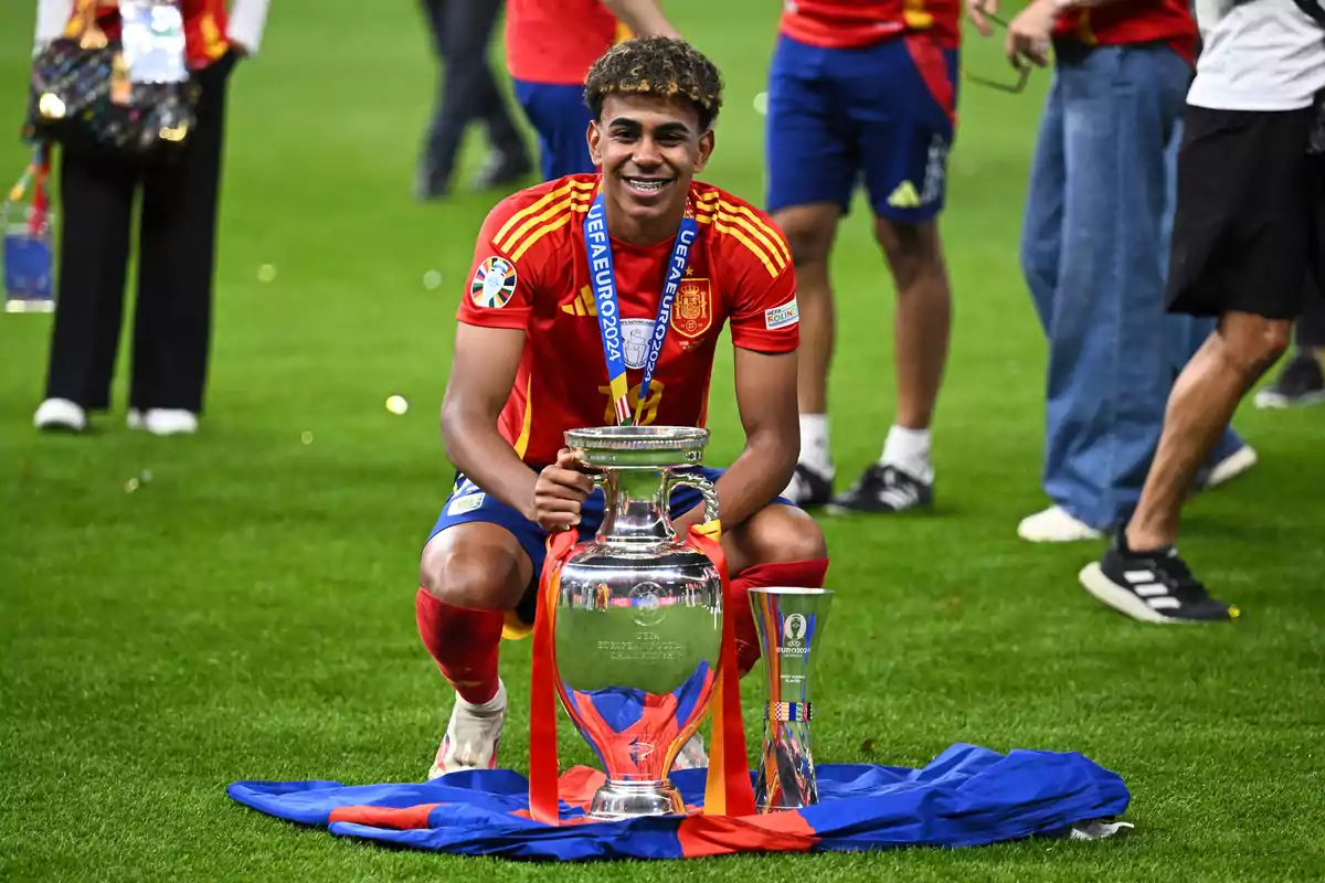 Un jugador de fútbol con el uniforme de la selección española sonríe mientras sostiene un trofeo grande y plateado en el campo de juego, con una medalla colgada al cuello y una bandera a sus pies.