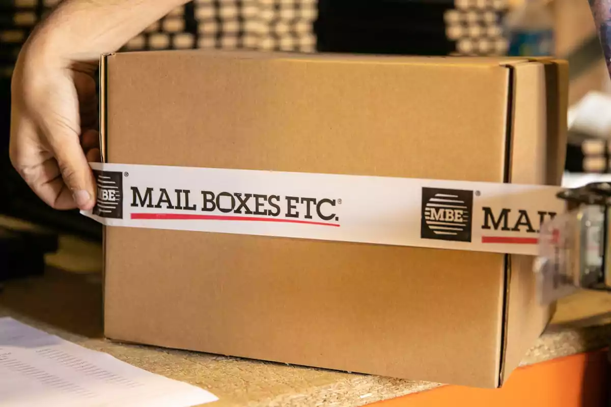 Mano sujetando una caja de cartón con cinta adhesiva de Mail Boxes Etc.