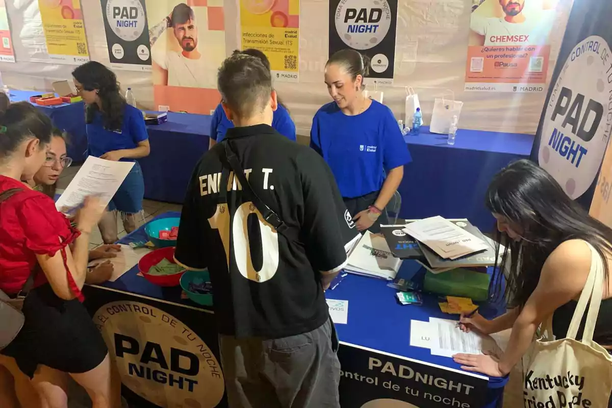 Personas interactuando en un stand informativo de PAD NIGHT, con carteles sobre salud sexual en el fondo.