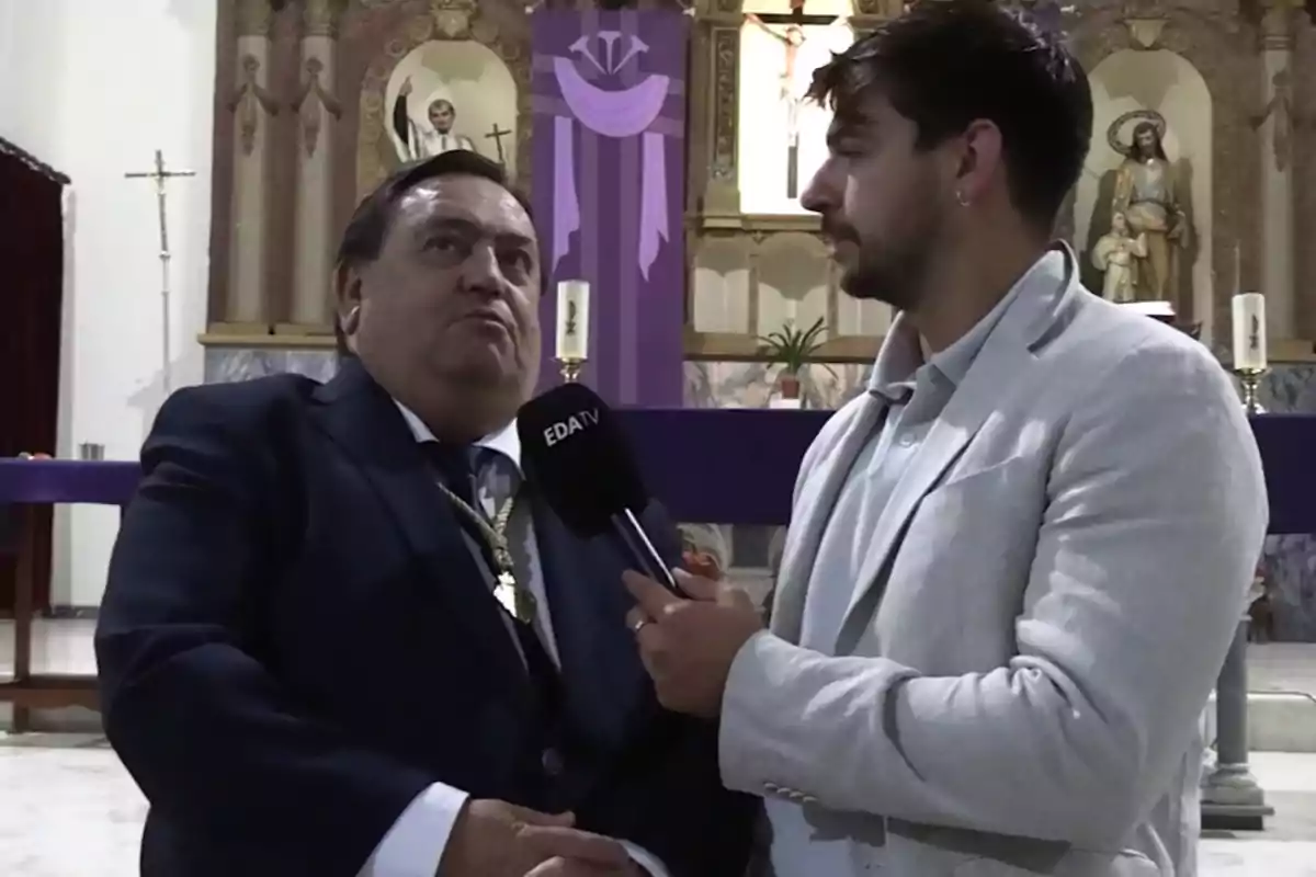 Un hombre con traje oscuro y medalla es entrevistado por otro hombre con chaqueta clara que sostiene un micrófono en el interior de una iglesia con decoraciones moradas y estatuas religiosas al fondo. Un hombre con traje oscuro y medalla es entrevistado por otro hombre con chaqueta clara que sostiene un micrófono en el interior de una iglesia con decoraciones moradas y estatuas religiosas al fondo.