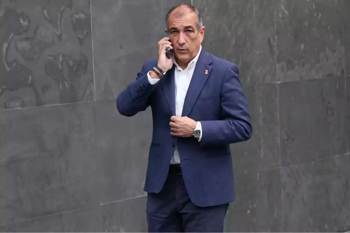 Hombre de traje azul hablando por teléfono móvil frente a una pared gris.