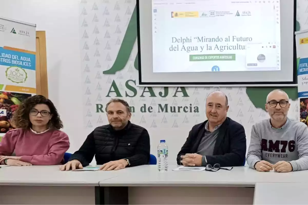 Cuatro personas sentadas en una mesa durante un evento de ASAJA Región de Murcia con un proyector mostrando el tema "Mirando al Futuro del Agua y la Agricultura".