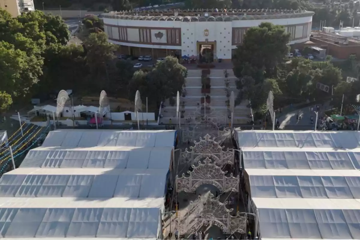 Vista aérea de una plaza de toros rodeada de árboles y estructuras cubiertas con carpas blancas, con una entrada decorada con arcos ornamentales y personas caminando por el acceso principal. Vista aérea de una plaza de toros rodeada de árboles y estructuras cubiertas con carpas blancas, con una entrada decorada con arcos ornamentales y personas caminando por el acceso principal.