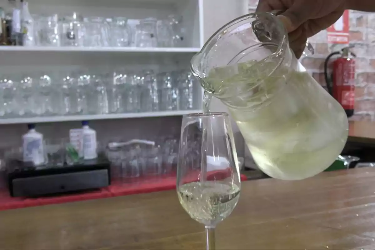 Una jarra de vidrio con hielo sirve una bebida clara en una copa sobre una barra, al fondo se ven estantes con vasos y copas de cristal. Una jarra de vidrio con hielo sirve una bebida clara en una copa sobre una barra, al fondo se ven estantes con vasos y copas de cristal.