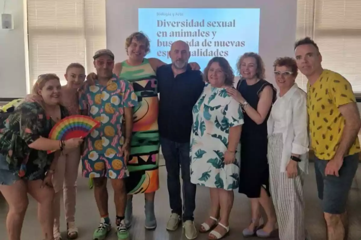 Grupo de personas sonrientes posando juntas en un aula con una pantalla al fondo que muestra el título Diversidad sexual en animales y búsqueda de nuevas especialidades, una persona sostiene un abanico con los colores del arcoíris. Grupo de personas sonrientes posando juntas en un aula con una pantalla al fondo que muestra el título Diversidad sexual en animales y búsqueda de nuevas especialidades, una persona sostiene un abanico con los colores del arcoíris.