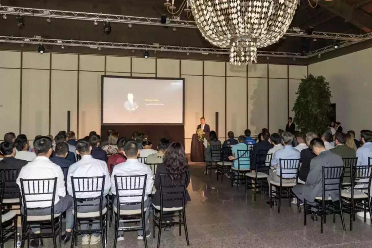Personas sentadas en un auditorio escuchando a un conferencista que está de pie junto a un podio frente a una pantalla grande en una sala elegante con una lámpara de araña
