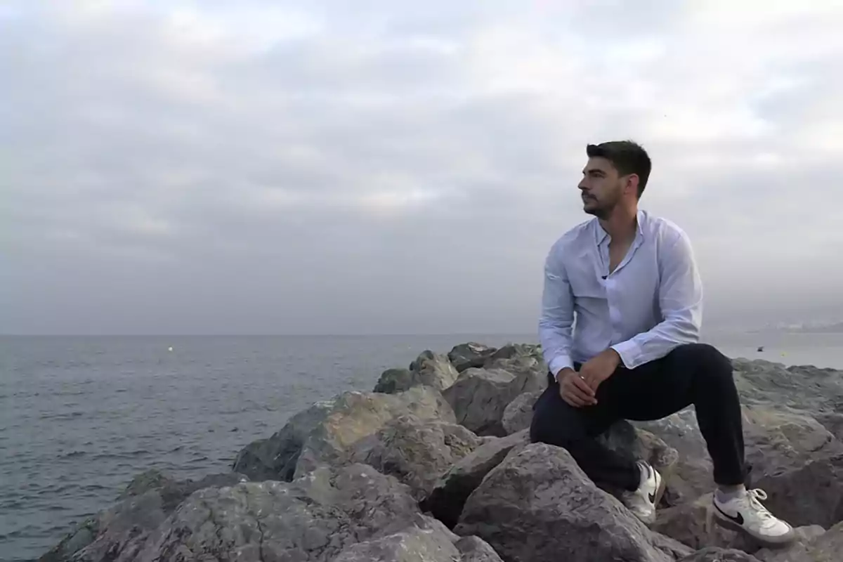Hombre sentado sobre rocas junto al mar mirando hacia el horizonte en un día nublado Hombre sentado sobre rocas junto al mar mirando hacia el horizonte en un día nublado
