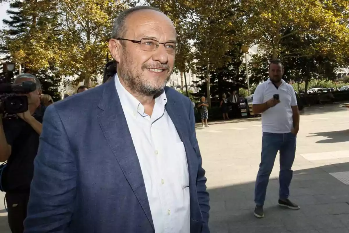 Hombre con gafas y barba sonríe mientras camina al aire libre con árboles y varias personas al fondo Hombre con gafas y barba sonríe mientras camina al aire libre con árboles y varias personas al fondo