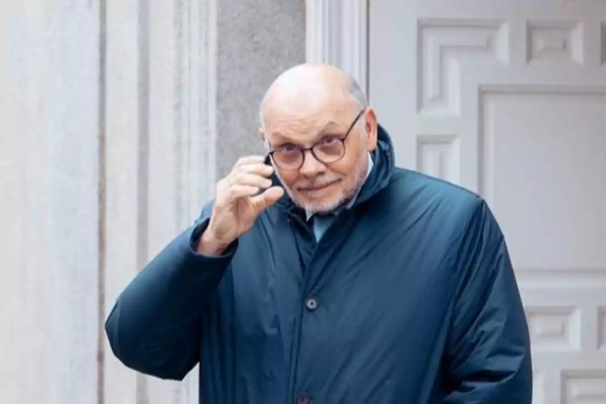 Hombre mayor con gafas y abrigo azul oscuro sonriendo y levantando la mano frente a una puerta blanca Hombre mayor con gafas y abrigo azul oscuro sonriendo y levantando la mano frente a una puerta blanca