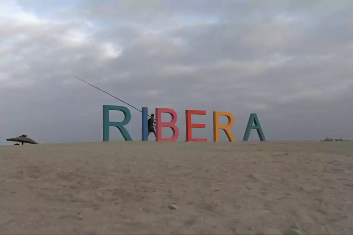 Letras coloridas que forman la palabra RIBERA sobre la arena con una persona pescando detrás de las letras y cielo nublado al fondo Letras coloridas que forman la palabra RIBERA sobre la arena con una persona pescando detrás de las letras y cielo nublado al fondo