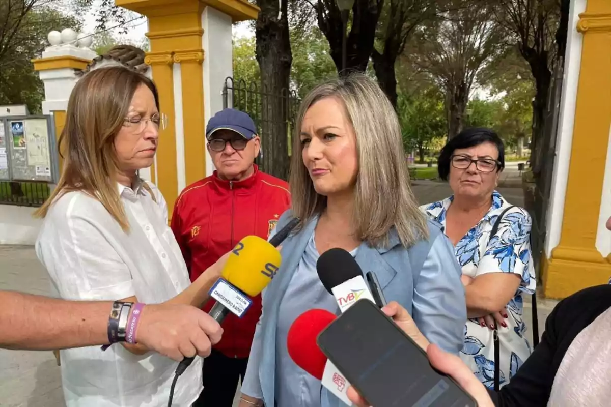 Una mujer está dando declaraciones a la prensa al aire libre mientras varias personas la rodean y varios micrófonos se acercan a ella Una mujer está dando declaraciones a la prensa al aire libre mientras varias personas la rodean y varios micrófonos se acercan a ella