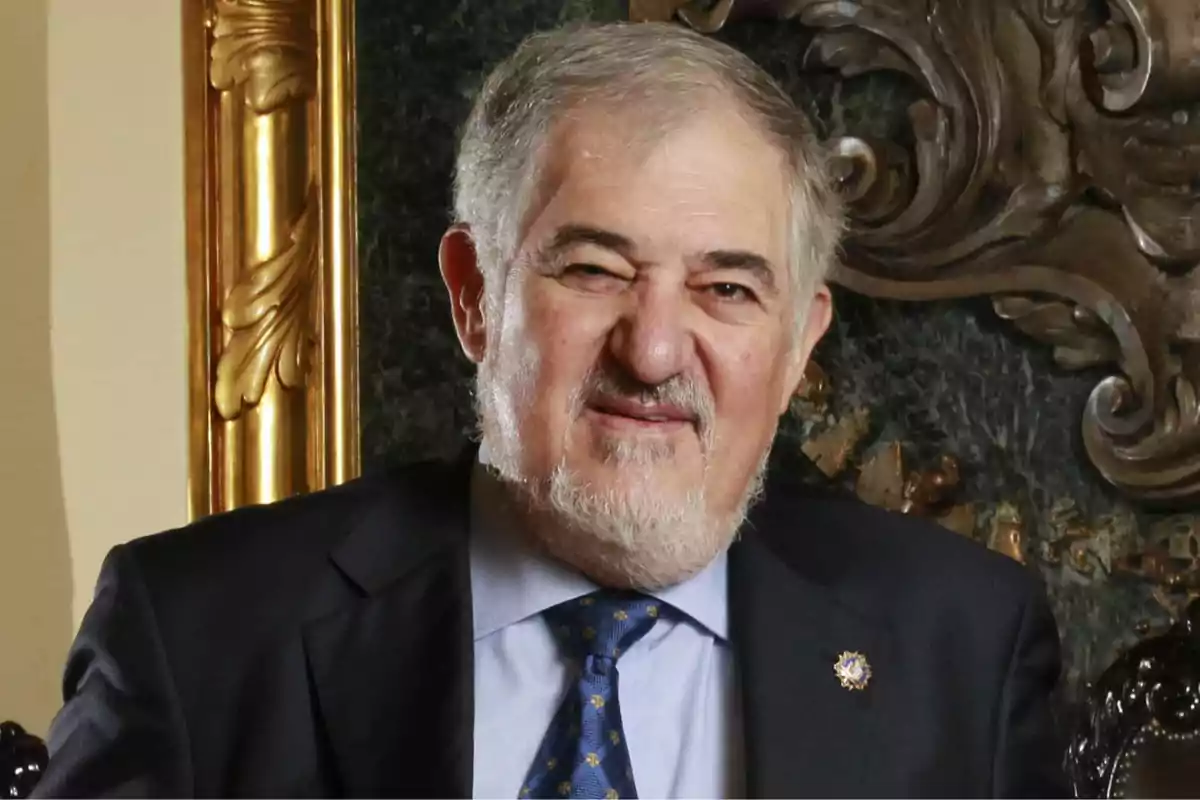 Hombre mayor de cabello canoso y barba corta vestido con traje oscuro y corbata azul sentado frente a un fondo decorativo elegante
