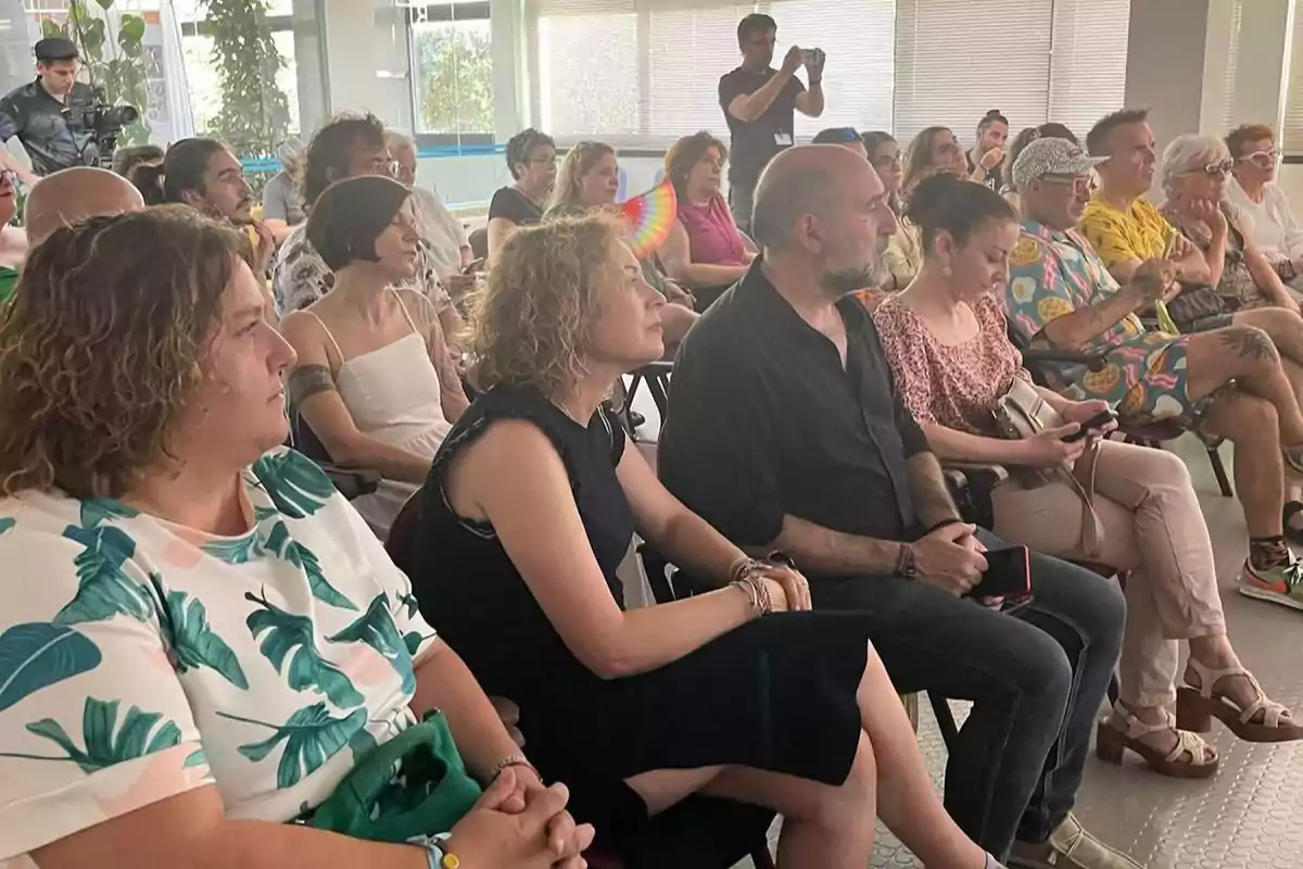 Personas sentadas en un auditorio prestando atención a un evento, algunas con ropa colorida y veraniega, mientras un hombre al fondo toma fotos con su celular. Personas sentadas en un auditorio prestando atención a un evento, algunas con ropa colorida y veraniega, mientras un hombre al fondo toma fotos con su celular.
