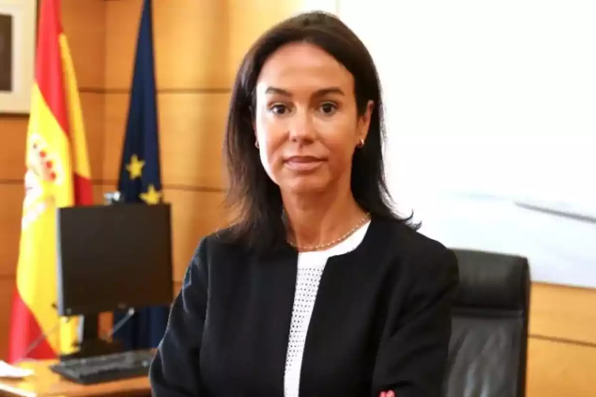 Mujer de cabello oscuro con chaqueta negra posando en una oficina con banderas de España y la Unión Europea al fondo Mujer de cabello oscuro con chaqueta negra posando en una oficina con banderas de España y la Unión Europea al fondo