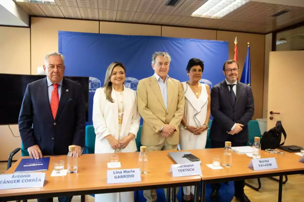 Cinco personas posan de pie detrás de una mesa en una sala de conferencias con fondo azul y banderas, sobre la mesa hay botellas de agua, vasos y carteles con nombres Cinco personas posan de pie detrás de una mesa en una sala de conferencias con fondo azul y banderas, sobre la mesa hay botellas de agua, vasos y carteles con nombres